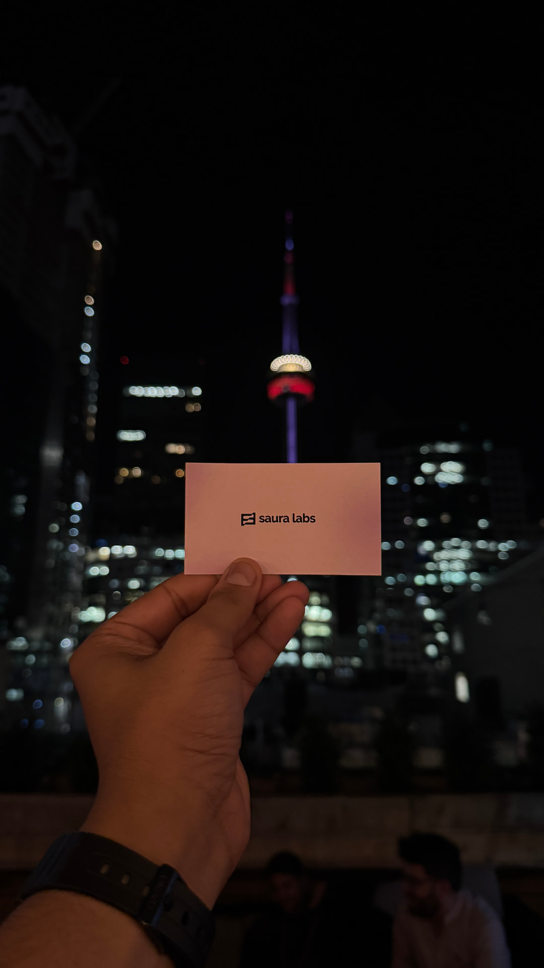 A Saura Labs business card held up against the CN Tower and the Toronto skyline at night.